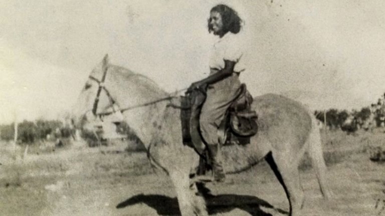 A black and white photograph of a smiling young woman atop a horse. 